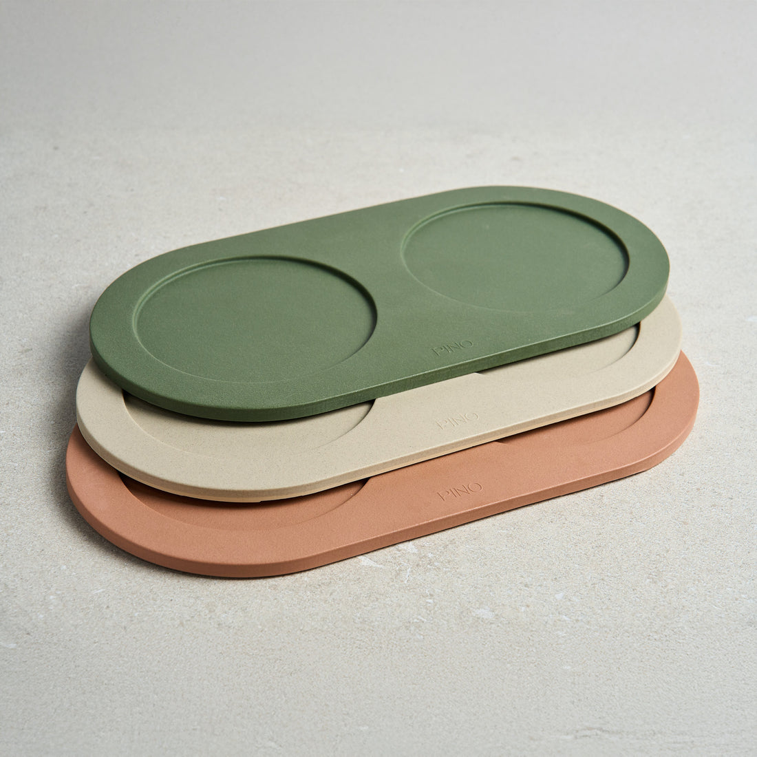 Three oval PINO trays stacked in green, cream and terracotta sandstone