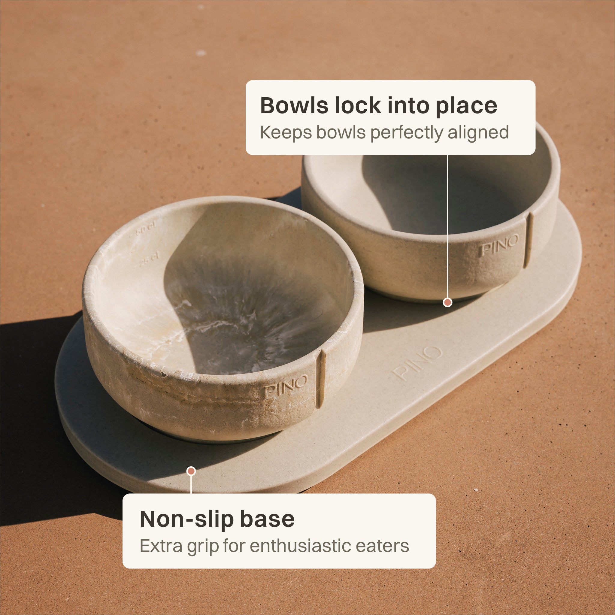 Two beige pet bowls on a matching non-slip base, shown on a warm brown surface with callout labels explaining alignment and grip.