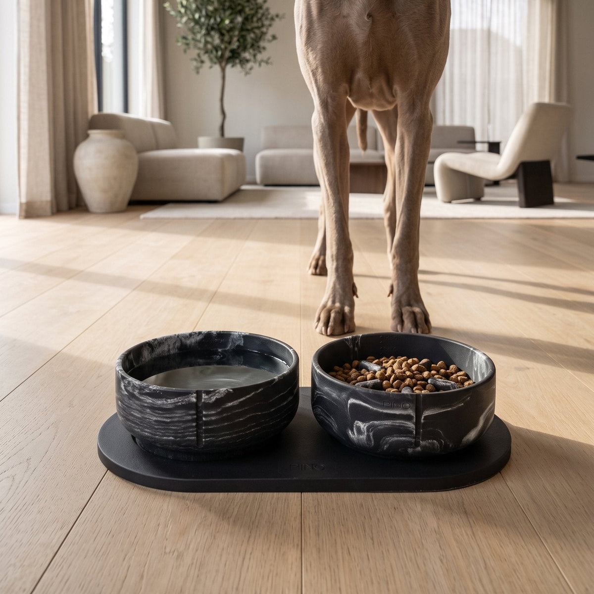 A weimaraner standing behind two marbled Black Panther bowls on a black Black Panther tray, one with water and one with kibble